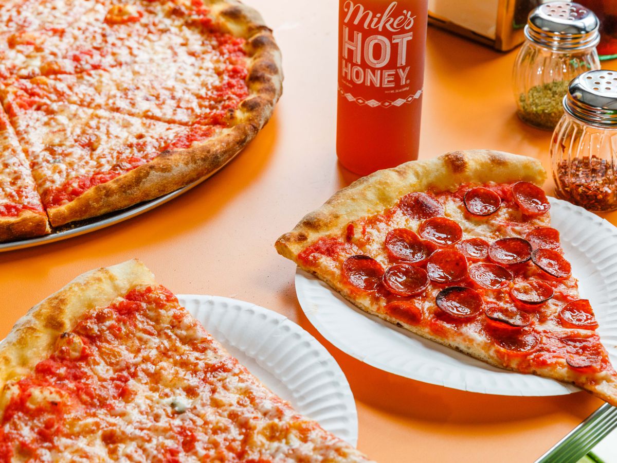 Pepperoni and cheese slices sit on an orange countertop at Paulie Gee’s slice shop, adjacent hot honey sauce and chile flakes