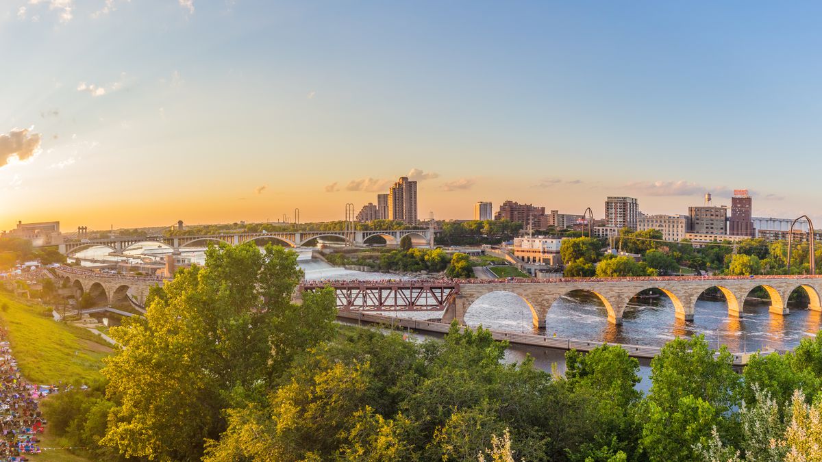 A wide landscape shot of the Mississippi River running through Minneapolis, An arched stone bridge runs across the river, connect both sides of the metropolis. The sun is setting and there are green trees in the foreground. 
