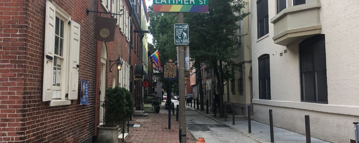 A street with a brick sidewalk. In the foreground is a lamp post with a street sign that says Latimer Street and a LGBTQ rainbow flag. On both sides of the street are businesses.
