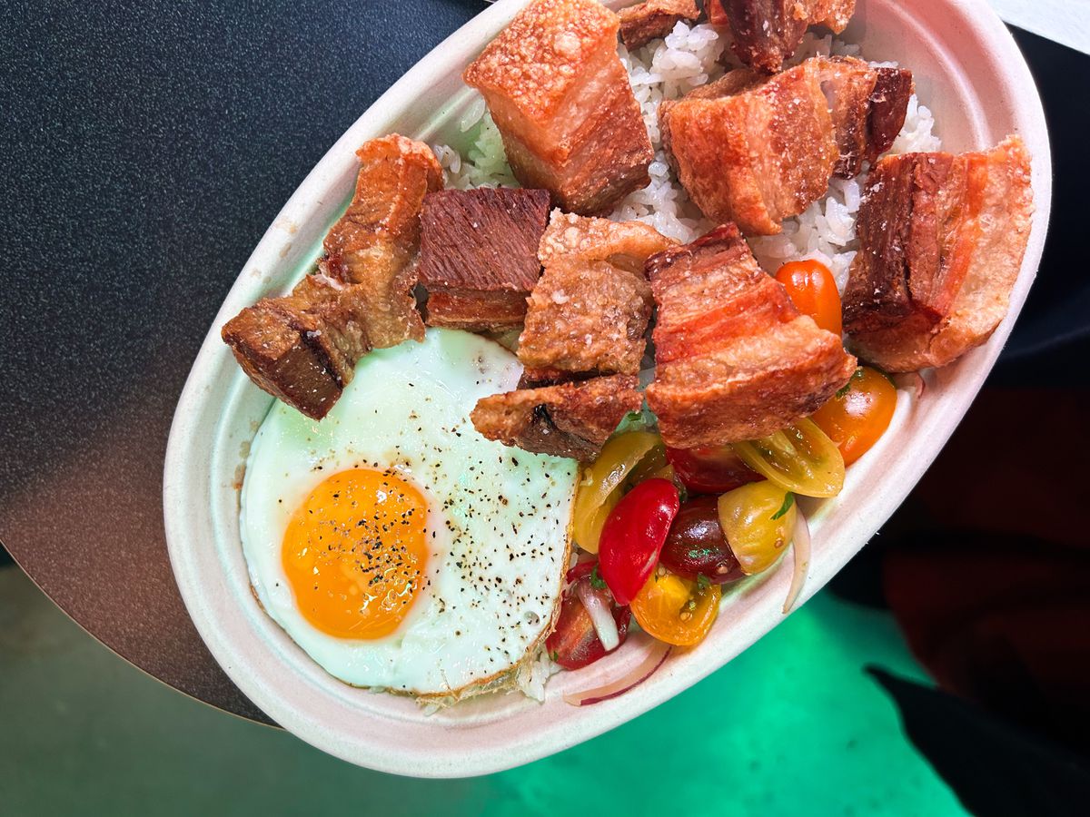 A crispy pork belly rice bowl at Sun Rice within the Moxy Hotel. The bowl comes with tomatoes and shallots, garlic rice, lechon kawali, and a sunny-side-up egg.