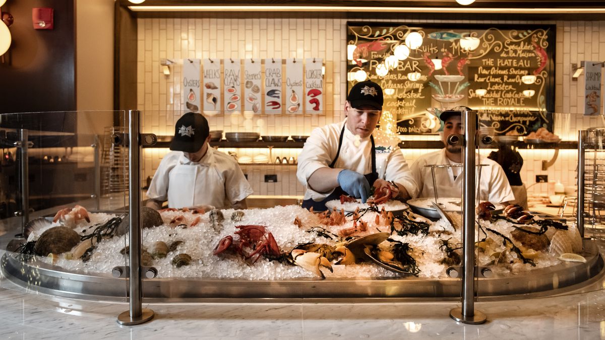Three chefs stand behind a clear partition working over an ice-covered raw bar with shellfish visible.