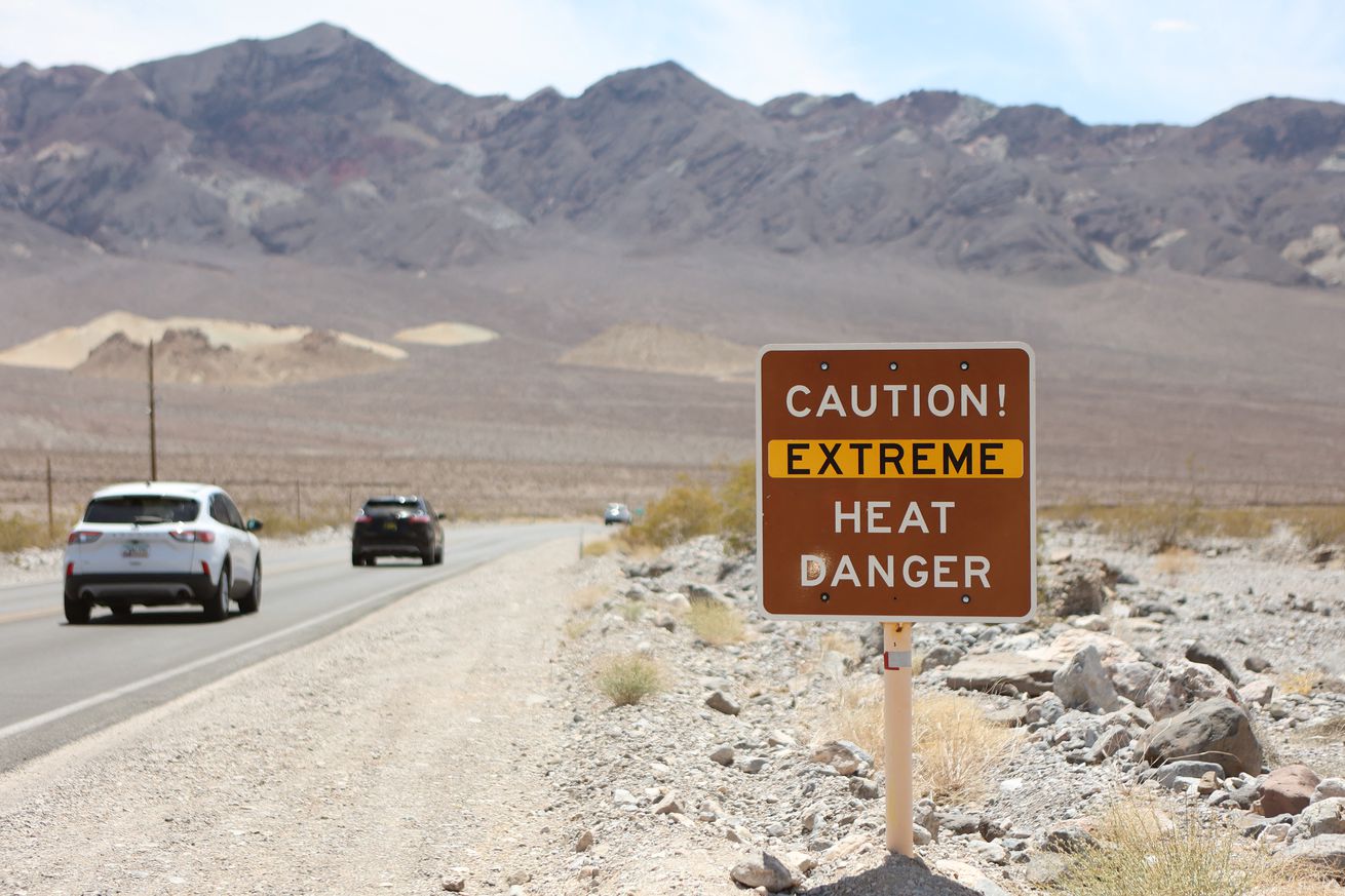 Cars drive on a desert road past a sign that says, “CAUTION! EXTREME HEAT DANGER”