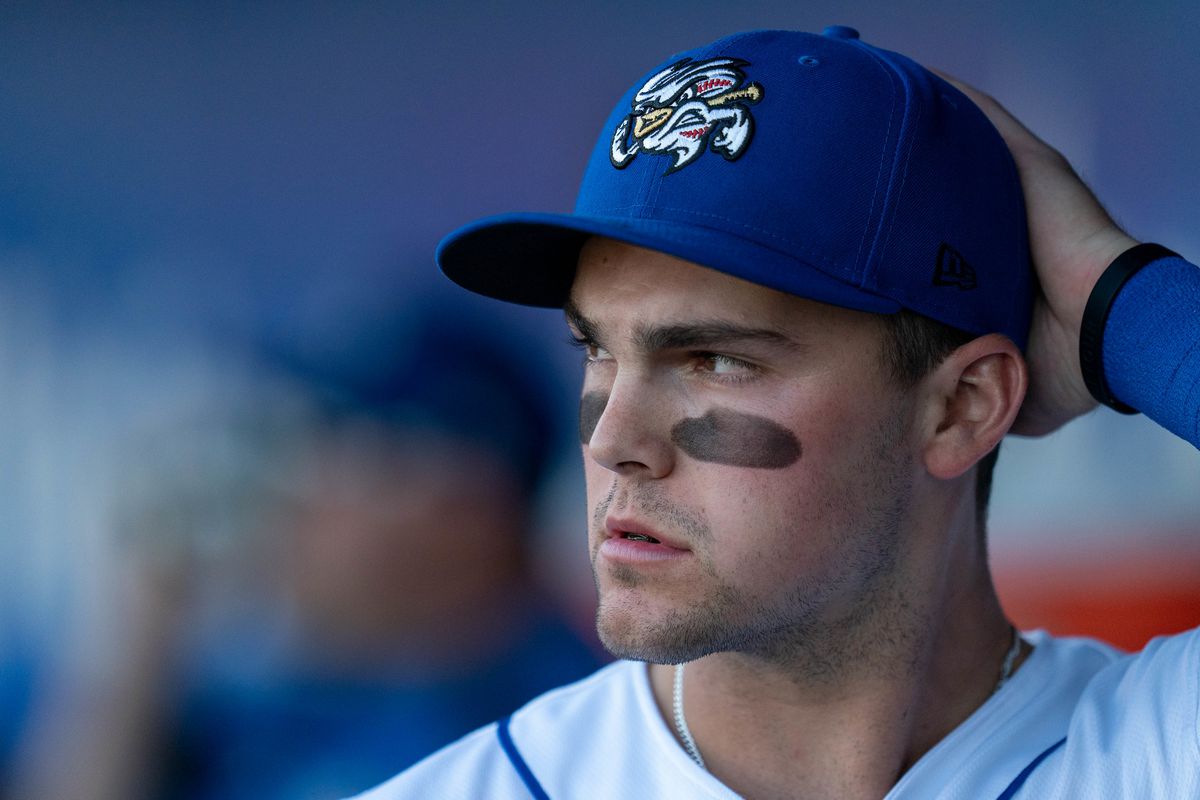 Closeup of a baseball player putting on a blue Omaha Storm Chasers baseball cap. He has eyeblack smeared under his eyes and a serious facial expression. 