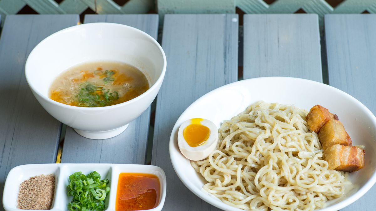 Two bowls, one of tonkotsu broth and another of noodles, sit on an slatted outdoor picnic table. Beside them, a tray of garnishes and colorful sauces.