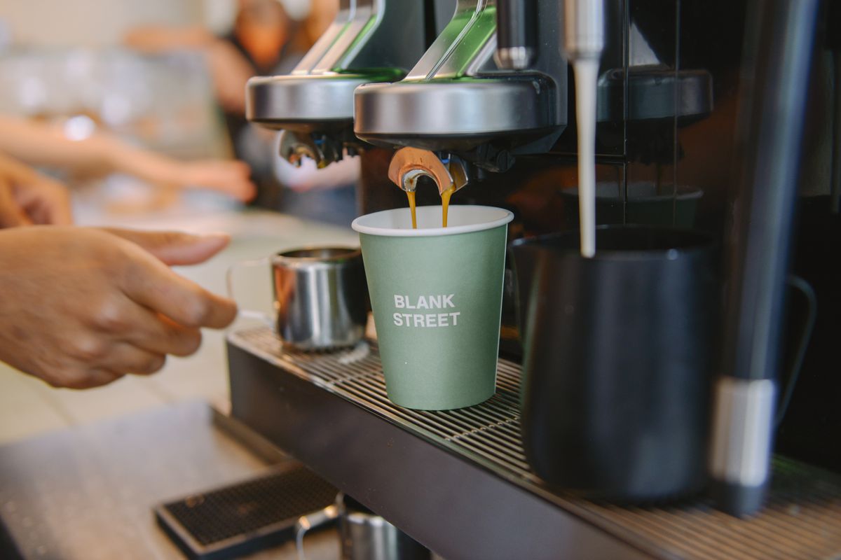 A green coffee cup with “Blank Street” written in white capital letters, under an espresso machine