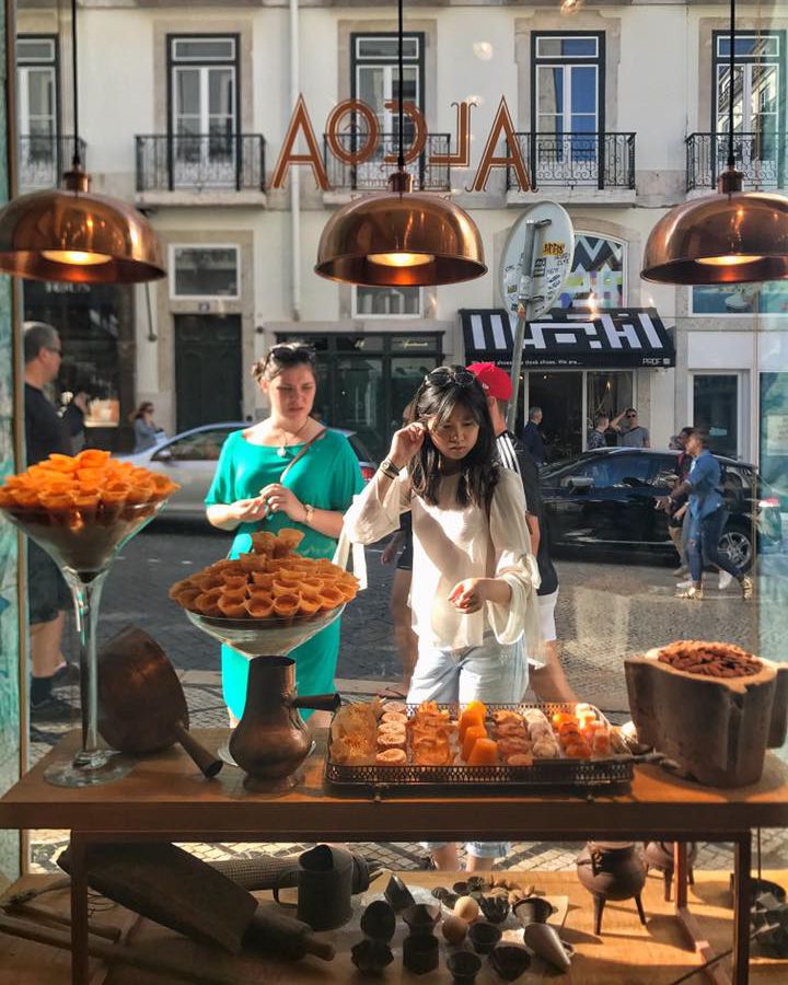 Two women stand outside in the sunshine beyond a shop window admiring displays of pastries sitting beneath bronze pendant lamps