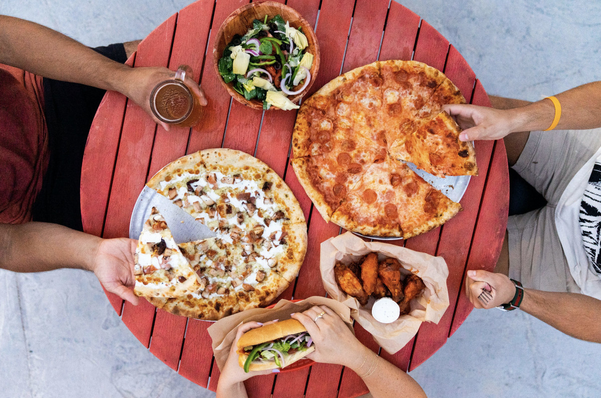 Three people seated around a round red table with two pizza, one sandwich, chicken wings, and a salad.