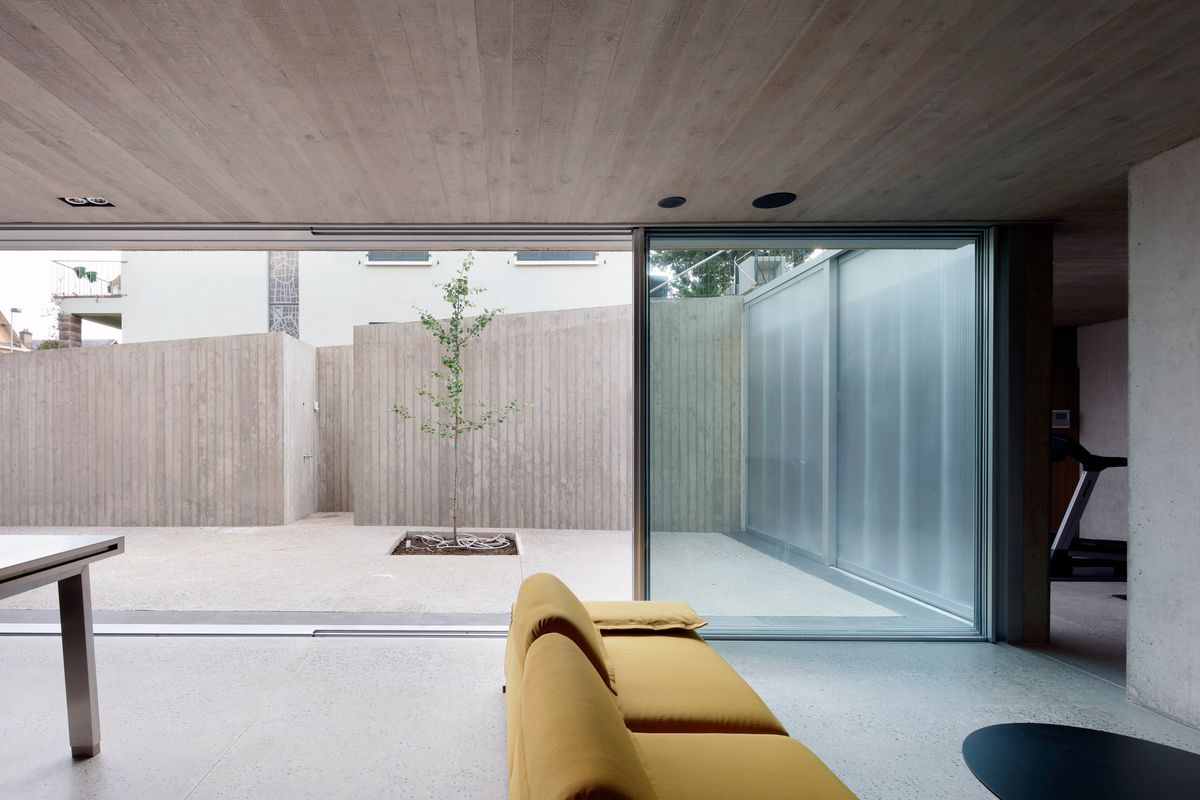 A living area with concrete floors, floor to ceiling windows overlooking a patio, and a yellow couch.