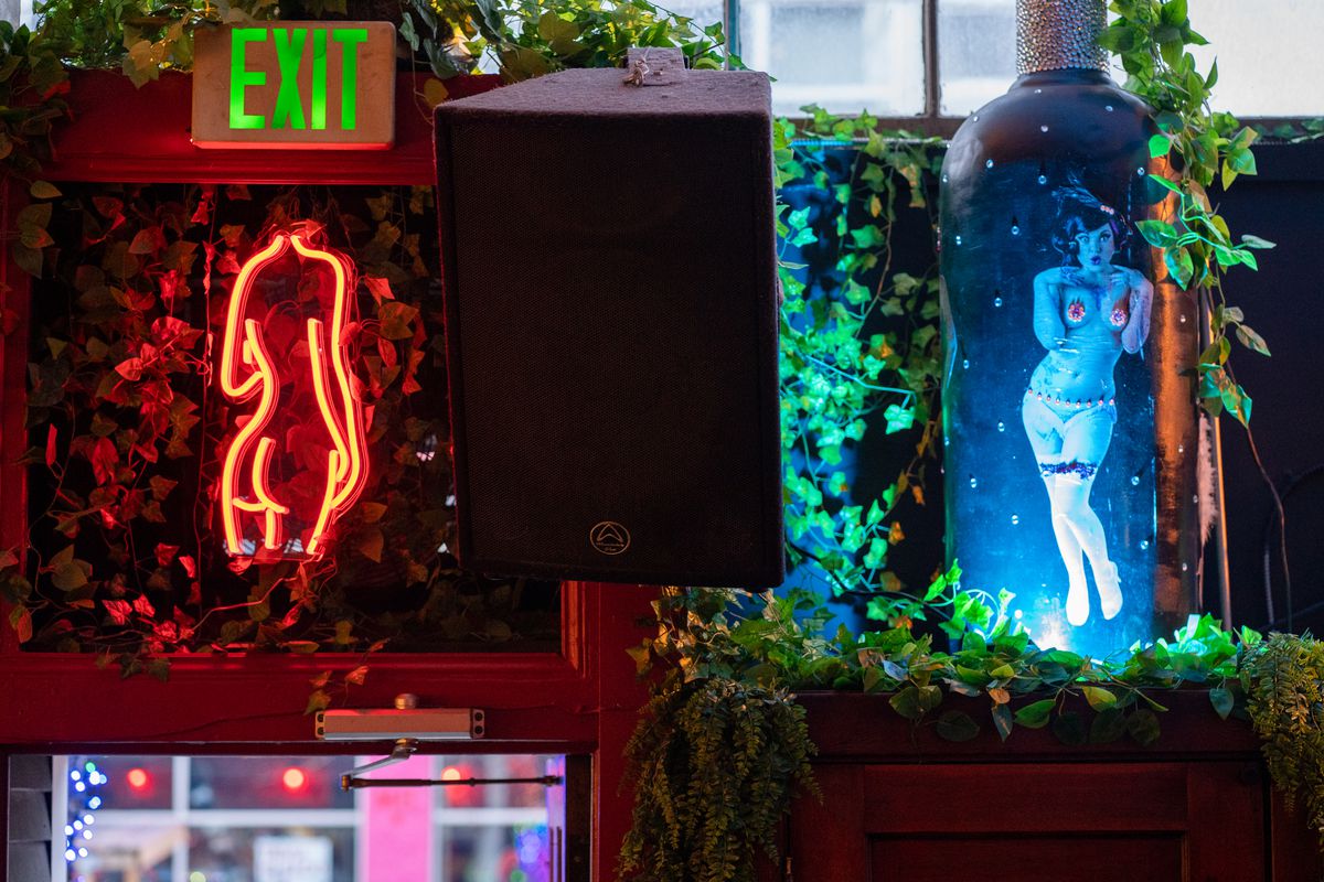 The view, from the inside, of the area above the front door of a bar, with an exit sign, a neon-light silhouette of a woman’s figure, and a large wine-bottle shaped objected decorated with a picture of a topless go-go dancer.