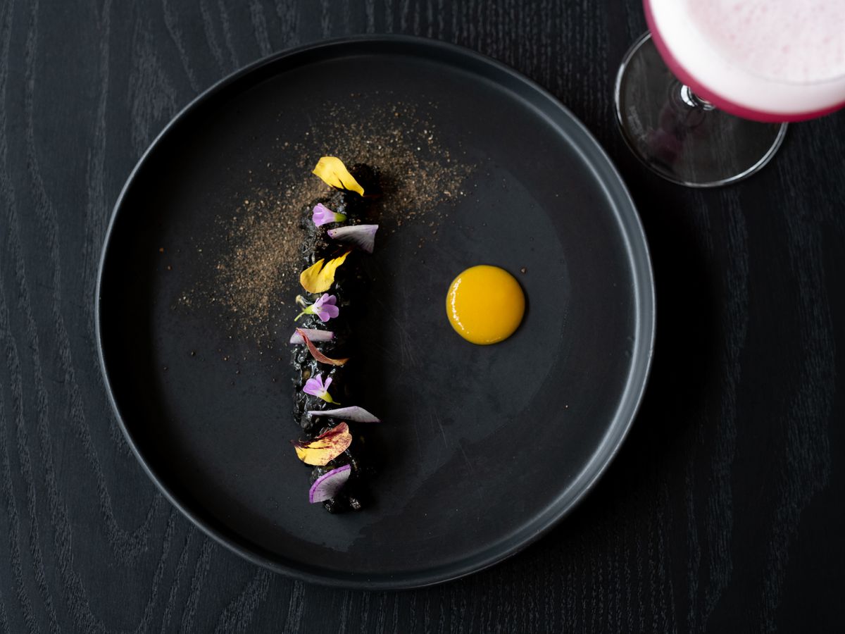 A stark black dish with a black central line of charred bits decorated with pops of yellow and lavender. A pool of golden sauce sits off to the side and a faint dusting of gold powder decorates the top left side of the plate.