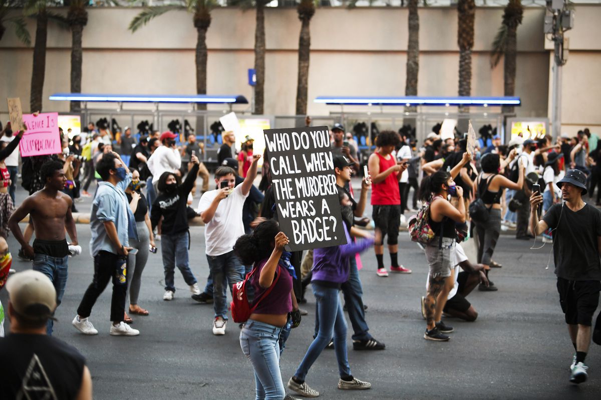 “Who do you call when the murderer wears a badge?” a black-and-white sign reads. Around the woman carrying it are socially distant knots of protesters.