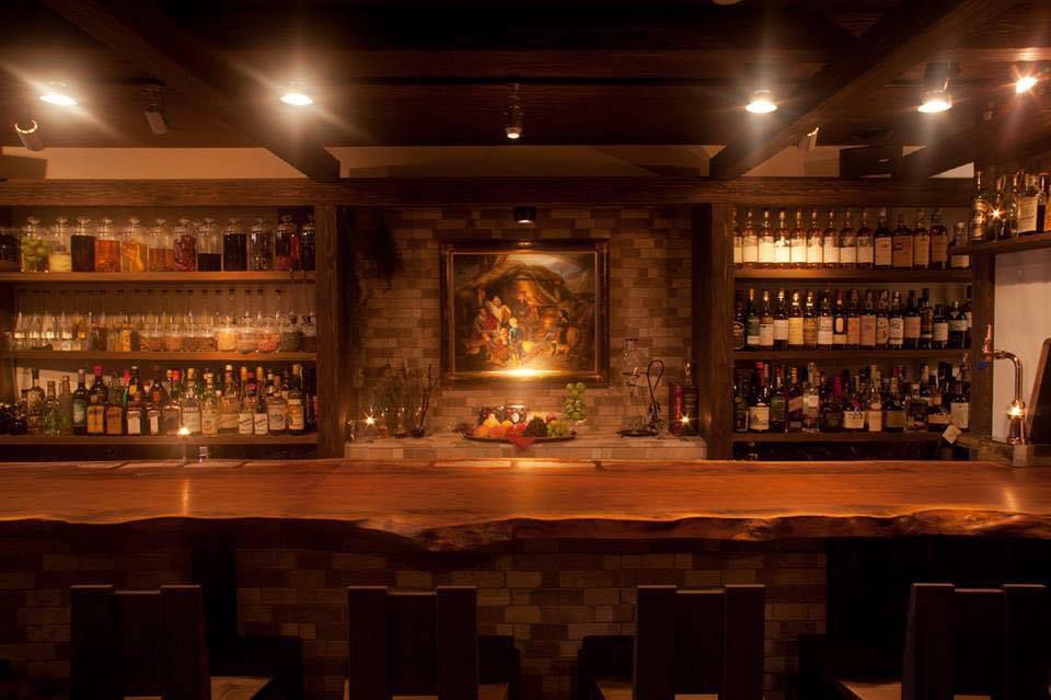 A darkened bar interior with shelves of backlit bottles, a dark wood bar, and a painting in the back.