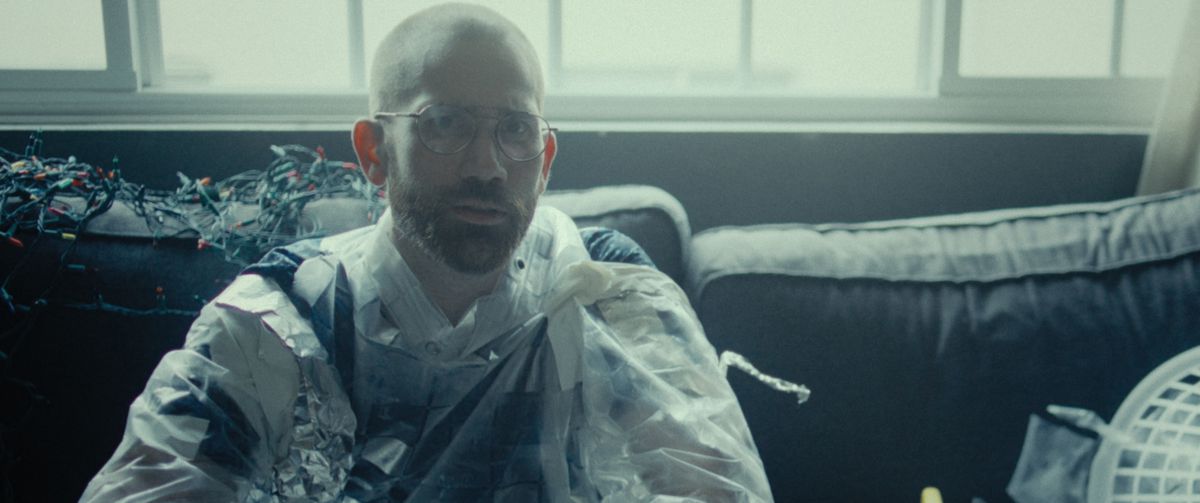 John Daniels (writer-director Aaron Moorhead) sits on a grimy couch in a dimly lit room, wearing a sloppy suit made out of masking tape, tin foil, and plastic sheeting, in Something In the Dirt