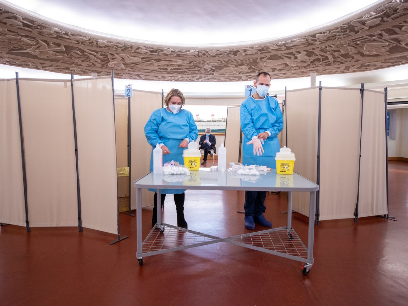 Two people stand at a table in protective gowns, gloves, and masks in preparation for distribution of a flu vaccine.