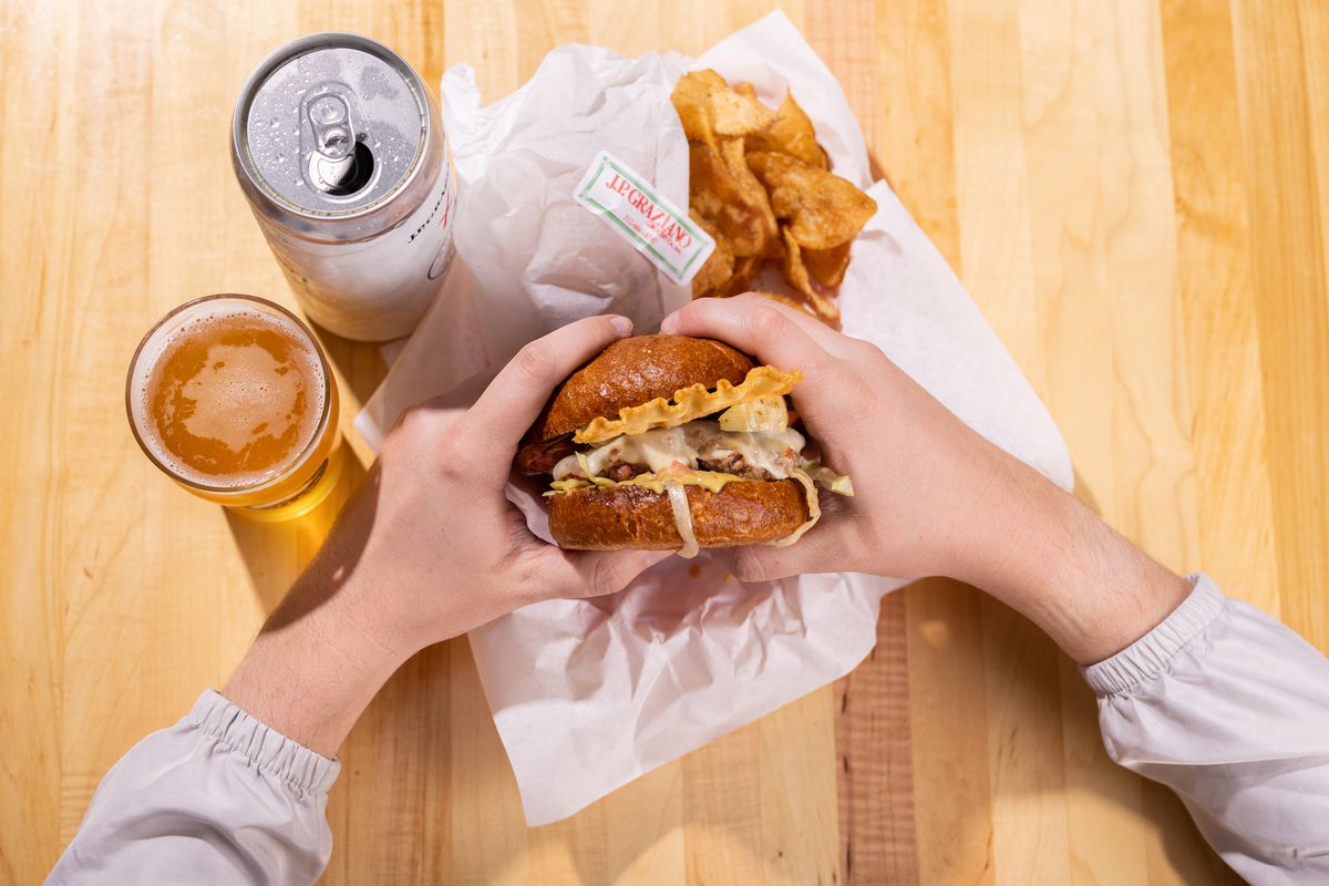 Two hands grabbing a cheese burger overhead with a beer in a glass and a can from a bird’s eye view.