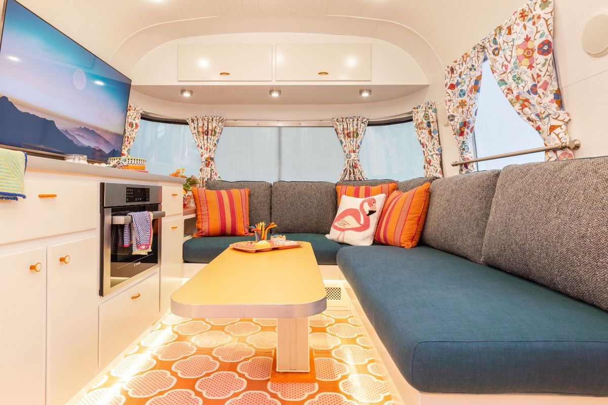 An interior view of a renovated Airstream travel trailer  has a teal blue bench seat, yellow table, and colorful print window coverings.