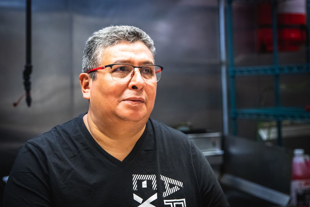 A person wearing glasses and a black T-shirt while inside a kitchen.