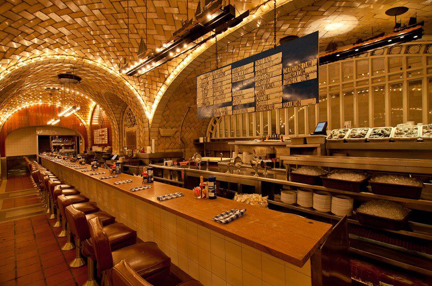 A long underground bar with backed bar stools is set up under an arching tunneled ceiling with yellow glowing lights.