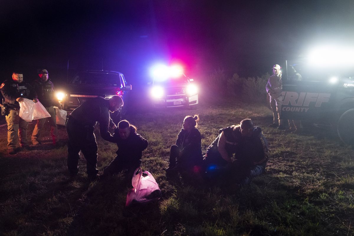 Flashing red and blue lights illuminate a nighttime scene of several migrants, including women and children, kneeling or sitting on the ground as police officers handcuff some of them.