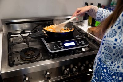A persons hand stirs food cooking in a pan on an indiction stove unit that is sitting on a gas stove.
