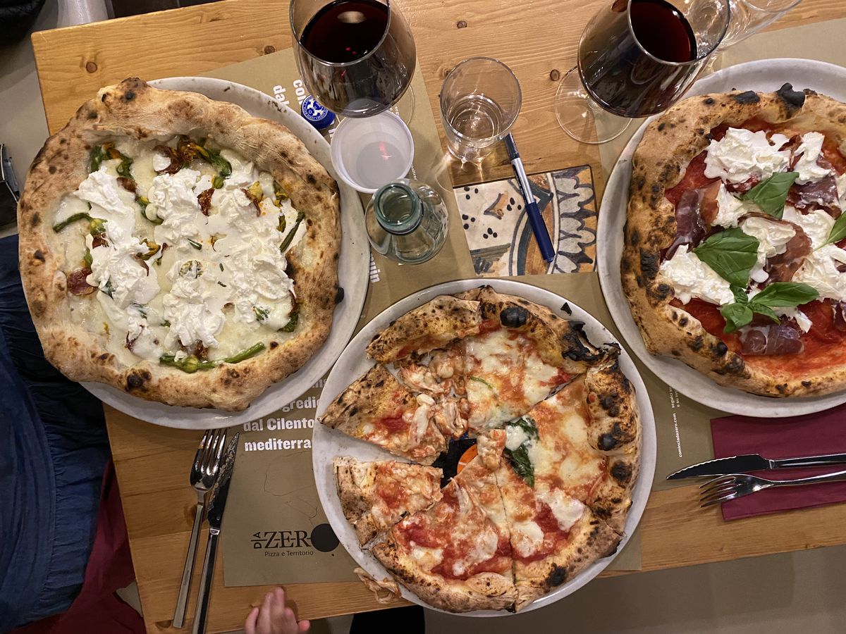 Three pizzas with various toppings in bright colors on a table beside glasses of red wine