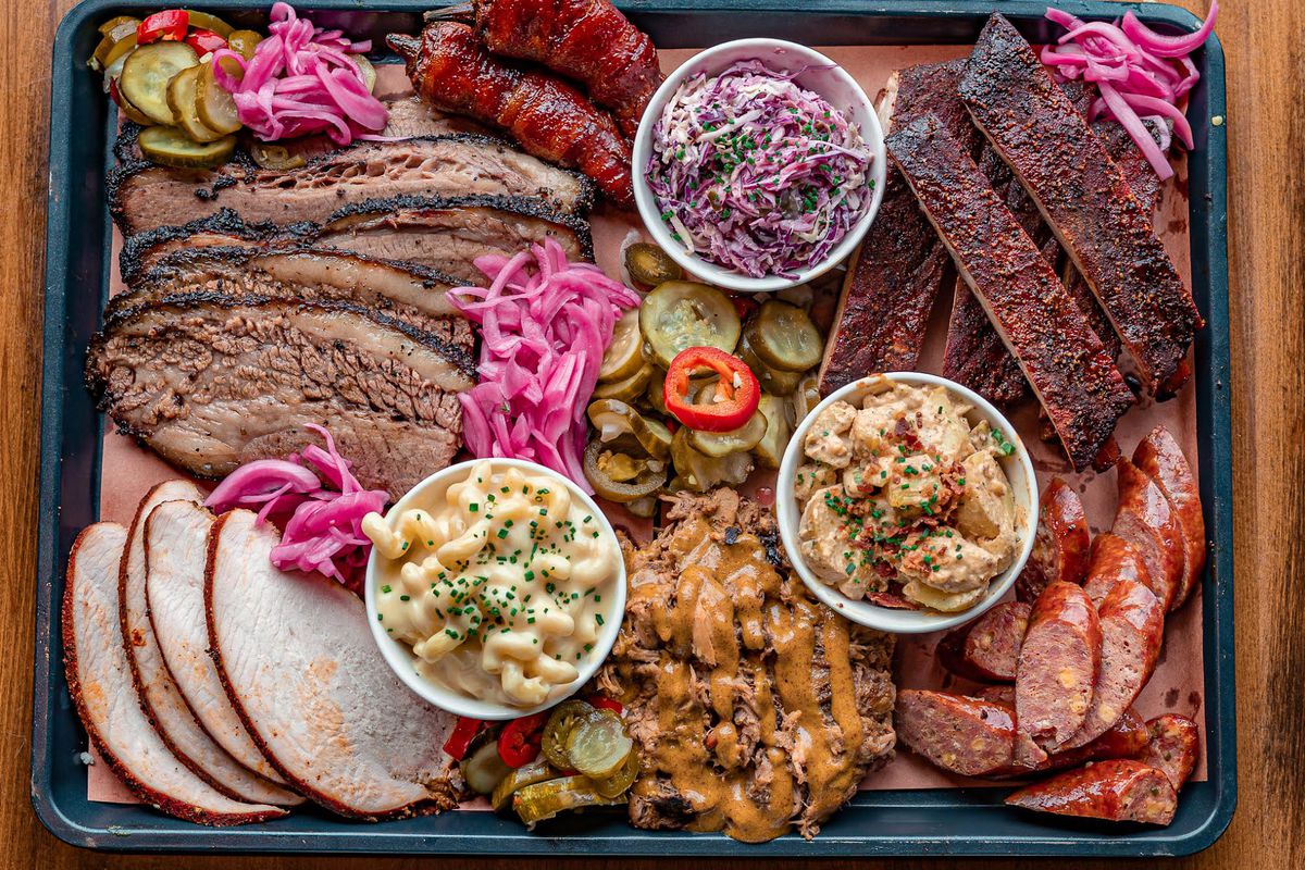 A plate of barbecue is loaded down with turkey, brisket, ribs, and sausage, with small containers of mac and cheese, cole slaw, and potato salad in the middle.