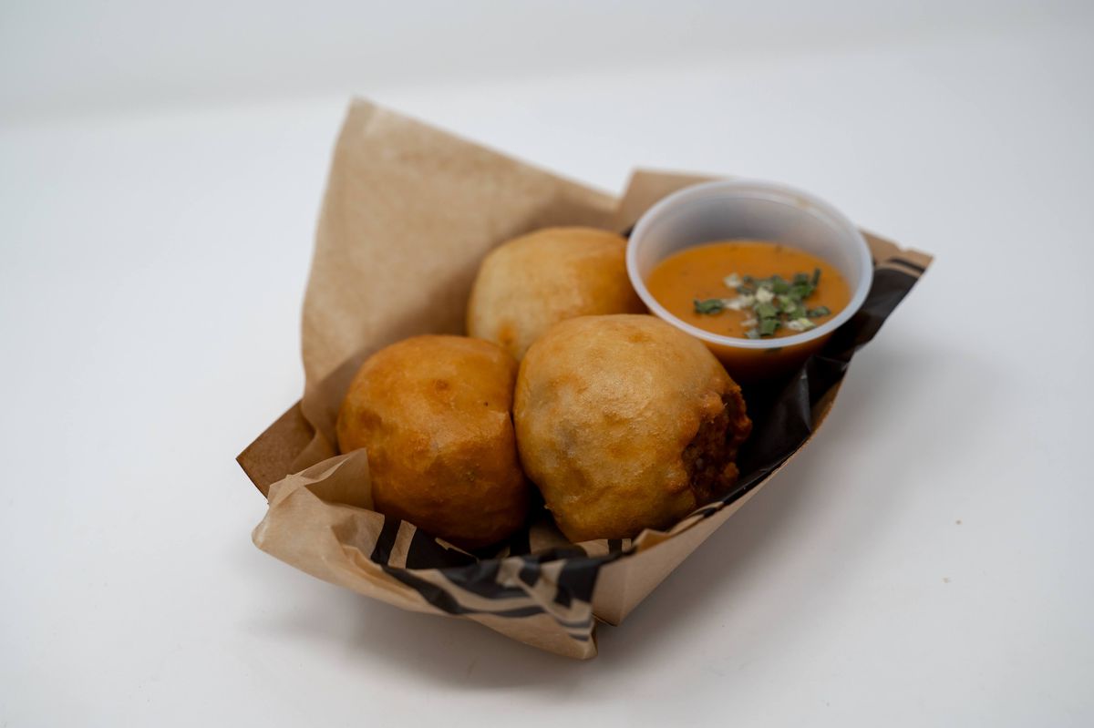 Three fried balls sit in a cardboard boat with brown paper. Next to them is a Creole dipping sauce that’s orange.
