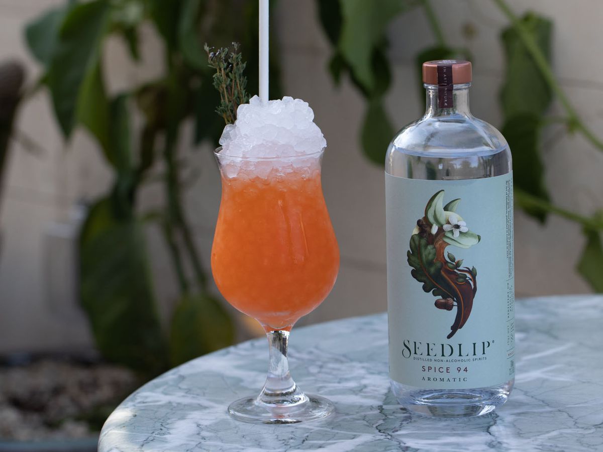 An orange, non-alcoholic cocktail filled with crushed ice, sits on a marble tabletop outside, next to a bottle of Seedlip distilled spirit. 
