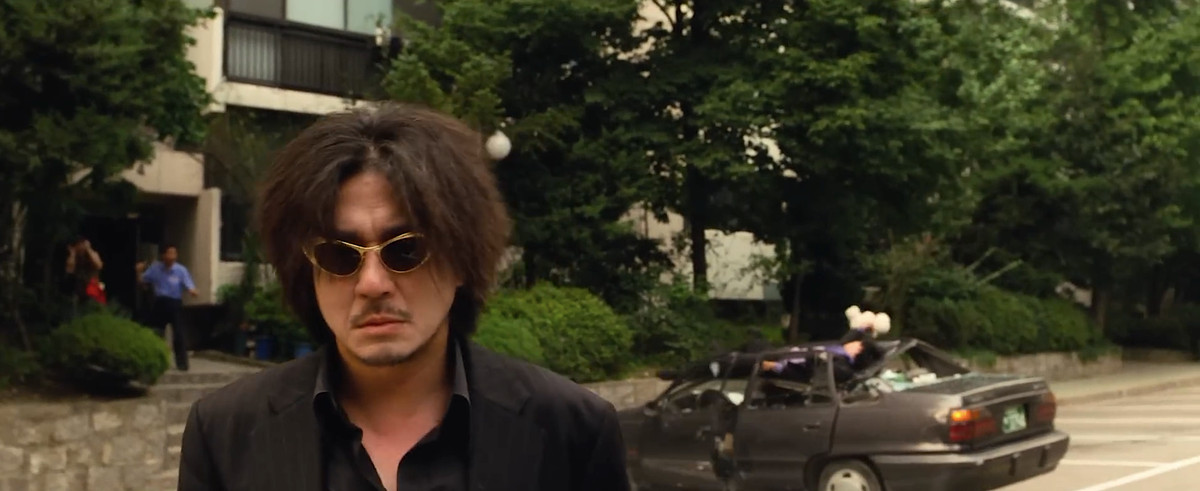 Oh Dae-su (Choi Min-sik), a shaggy-haired man wearing sunglasses and a black shirt, stands on the street looking expressionless, in front of a car that’s been crushed by a falling human body in Park Chan-wook’s Oldboy