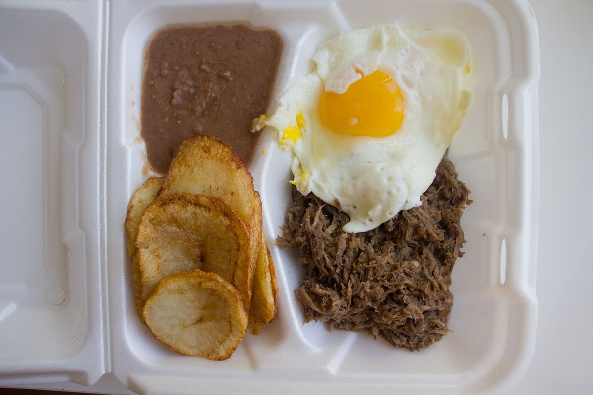 A container of barbacoa meat, sunny-side up fried egg, sliced potatoes, and refried beans. 