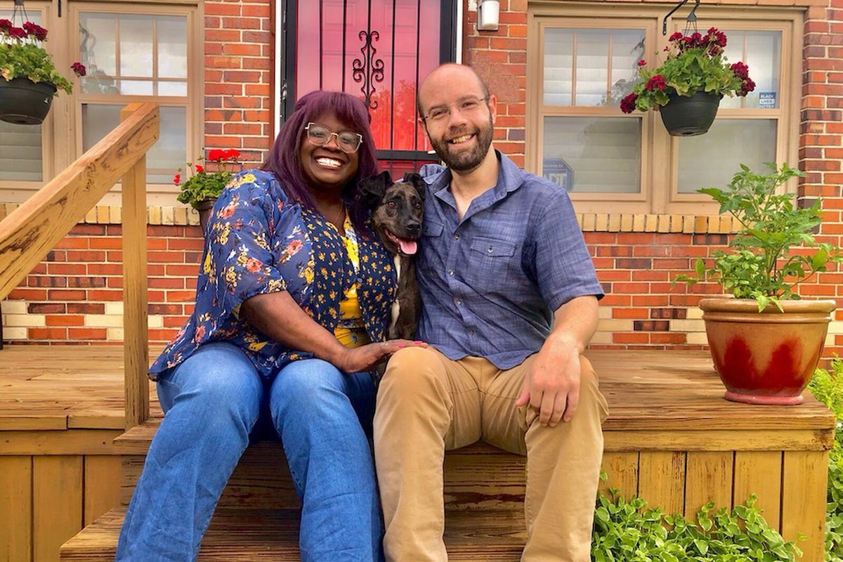A man and a woman sit on a wooden porch. They are smiling at the camera. There is a dog between them with its tongue out.