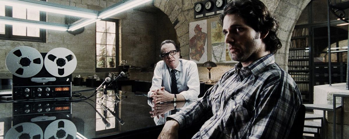 Avner (Eric Bana) sits with an older Mossad agent in a white shirt and glasses (Geoffrey Rush) while listening to a reel to reel tape recording in a spy headquarter basement in 2005’s Munich