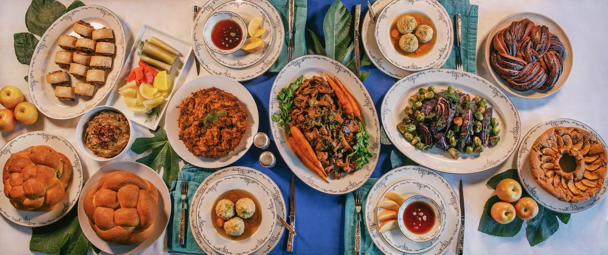 A blue and white table cloth topped with vintage china and traditional Rosh Hashanah dishes, including: a rugelach platter, three braided loaves of challah, matzoh ball soup, mushroom brisket, cider-braised cabbage, apples and honey, and apple cake (flanked by three whole apples).