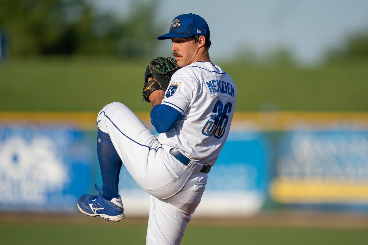 Upper body photo of a baseball pitcher in a white uniform and blue cap, with a neatly twirled mustache, rearing back to throw a pitch.