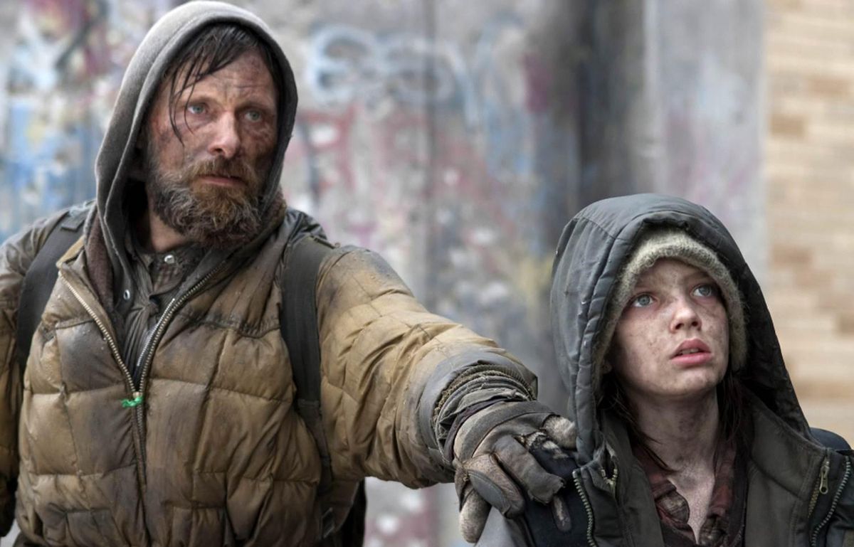 A disheveled man (Viggo Mortensen) in a dirty coat places his hand on the shoulder of a young boy (Kodi Smit-McPhee) looking up at something off-screen in The Road.