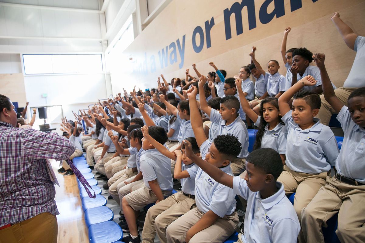 A large group of students, wearing light blue shirts and khaki pants, raise their hands seated on bleachers while educators stand in front of them.