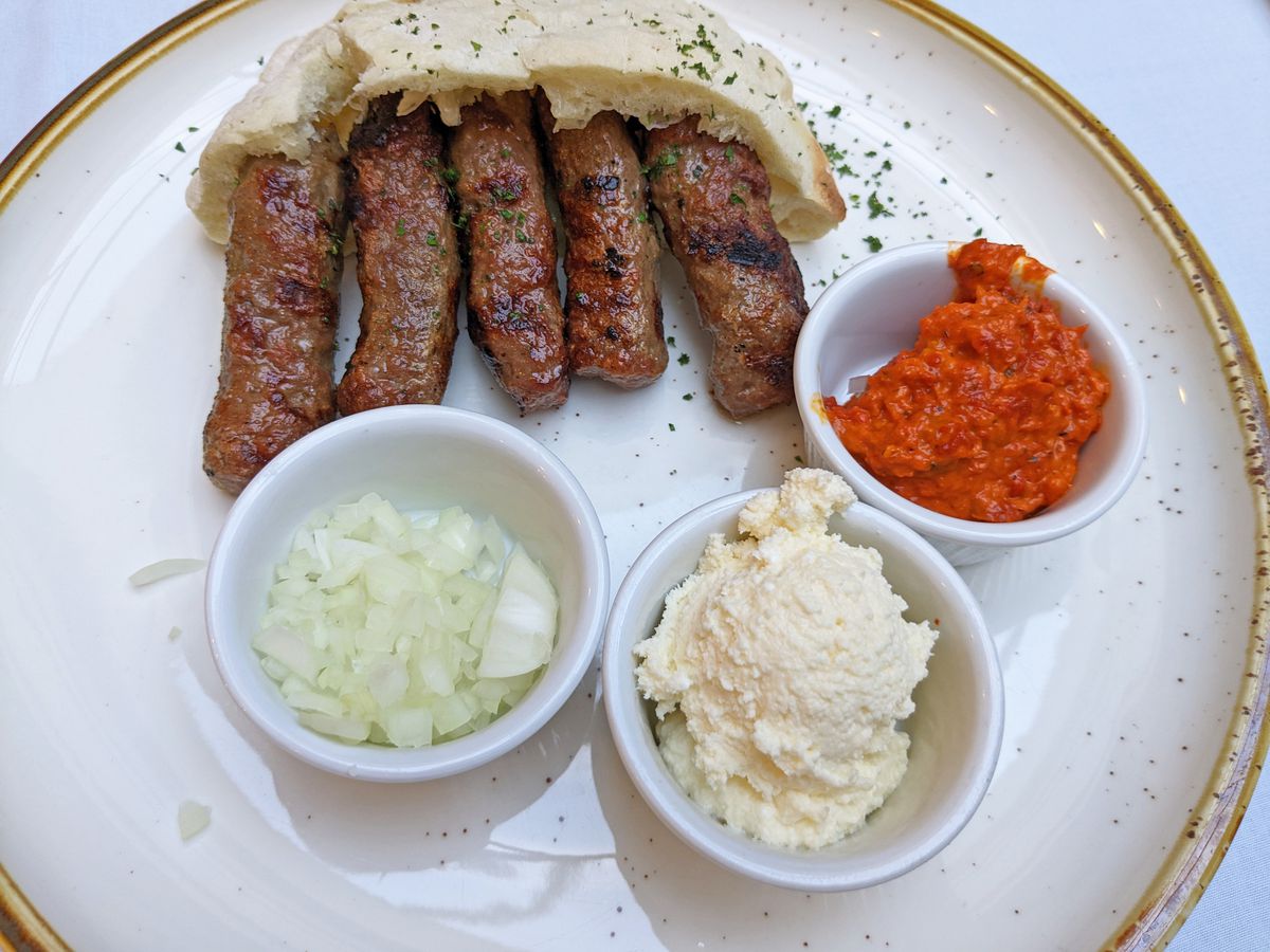 A plate with colored small dishes of onions, red paste, whitish paste, along with five sausages and a round bread.