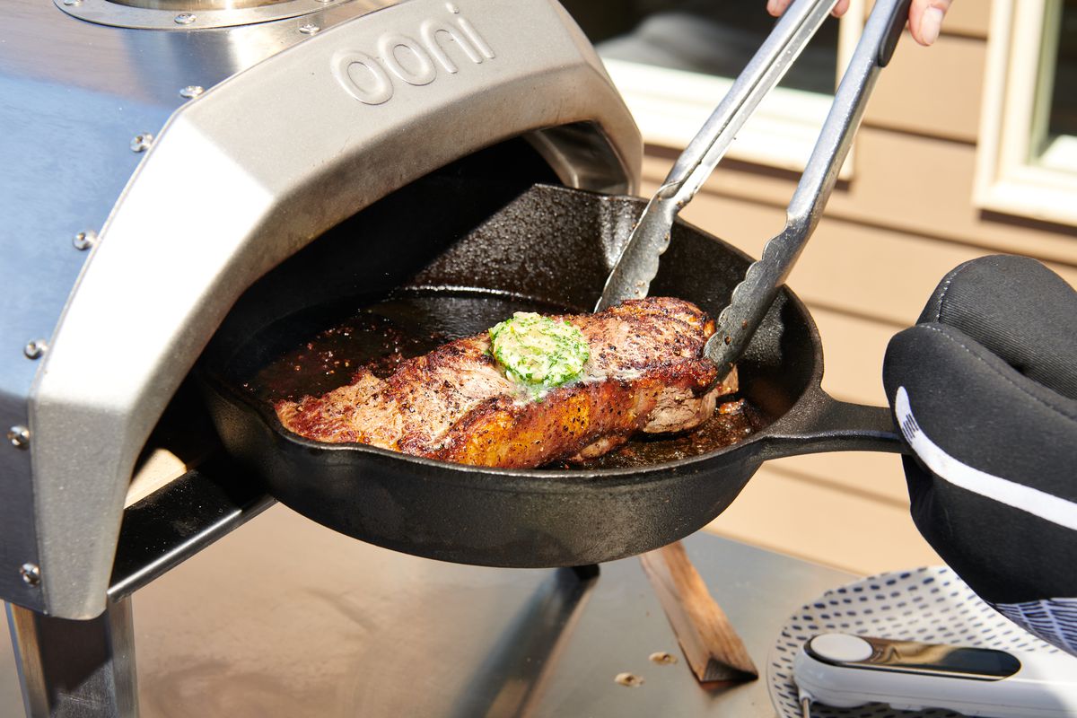 A cast iron skillet holding a steak with compound butter on top is pulled out of a pizza oven and adjusted with tongs.