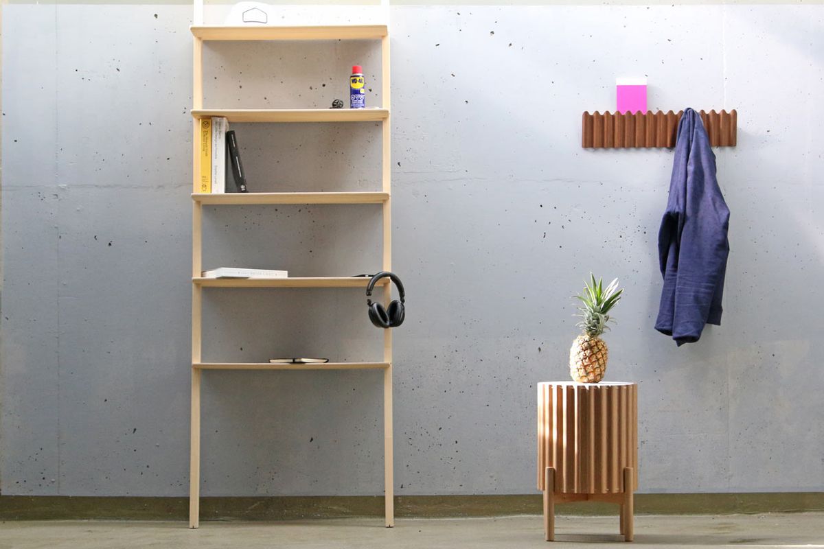 Shot of a leaning shelf, round side table, and coat rack all made of corrugated plywood set against a concrete wall in a plain room. 
