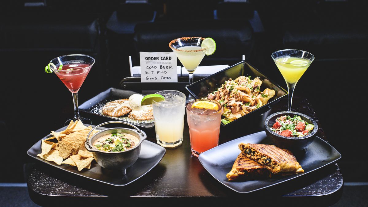 Several cocktails and plates of comfort food are arranged on a dark background, with an order card reading “cold beer, hot food, good times.”