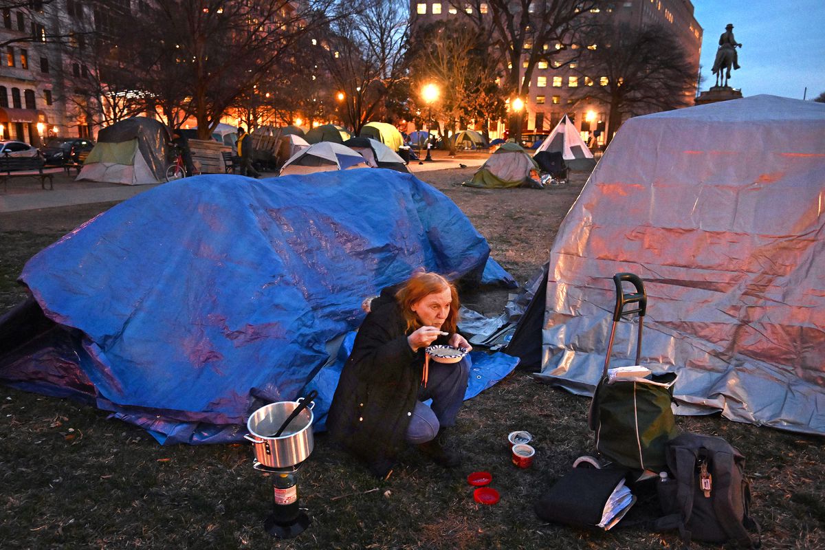 A woman sitting outside a tent eating soup with other tents in a homeless encampment in the background, and lit-up city buildings behind that.
