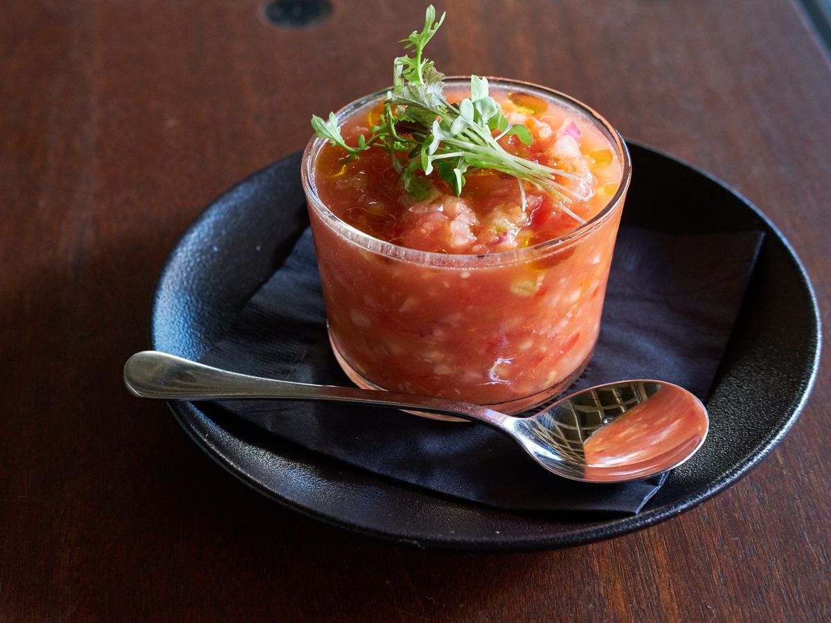 A clear cup of red gazpacho topped with fresh greens, on top of a black plate next to a small spoon.