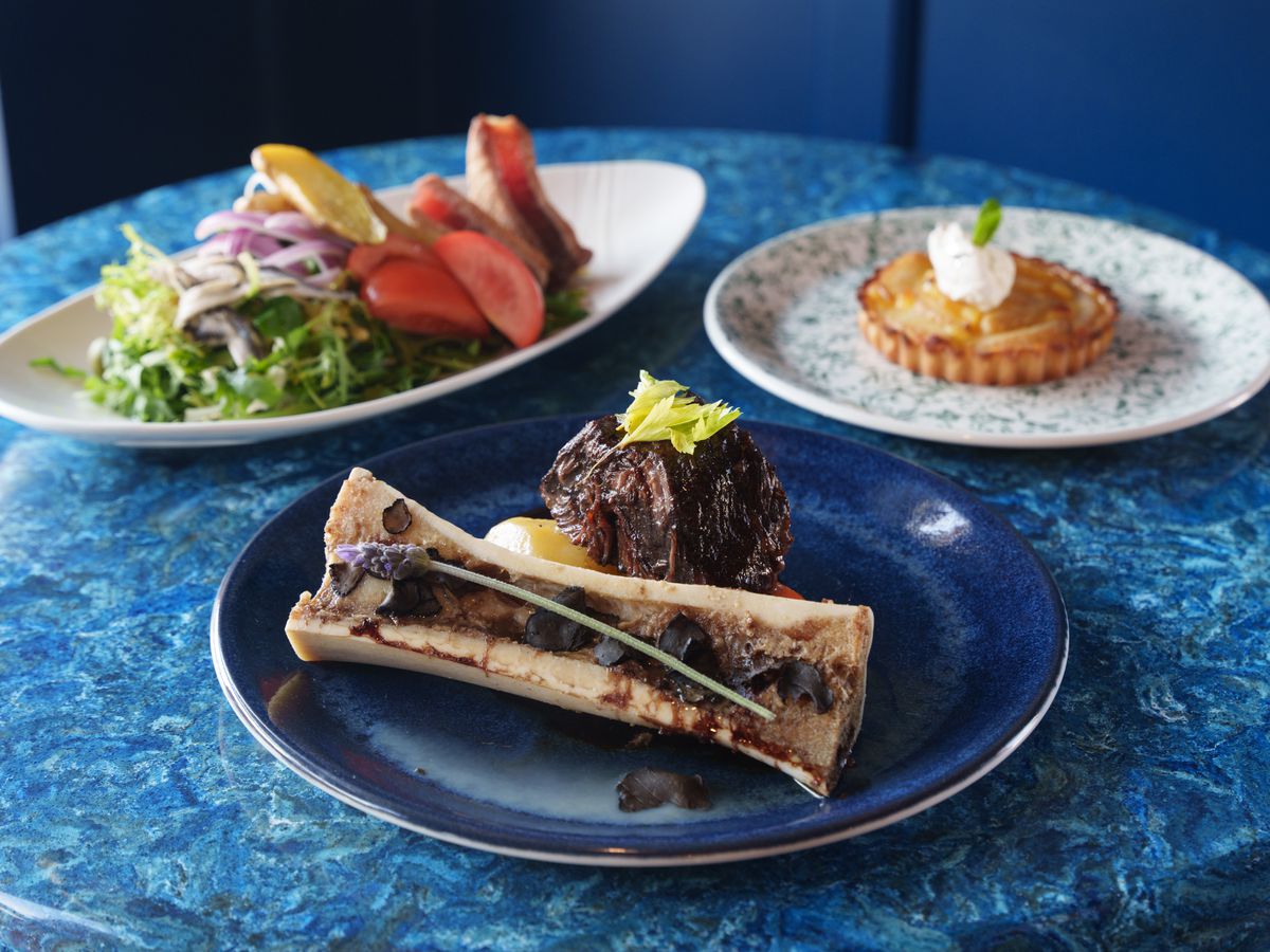 Three dishes on a blue table: A bone marrow beef dish (center), a Nicoise salad (left) and a pear tart (right).