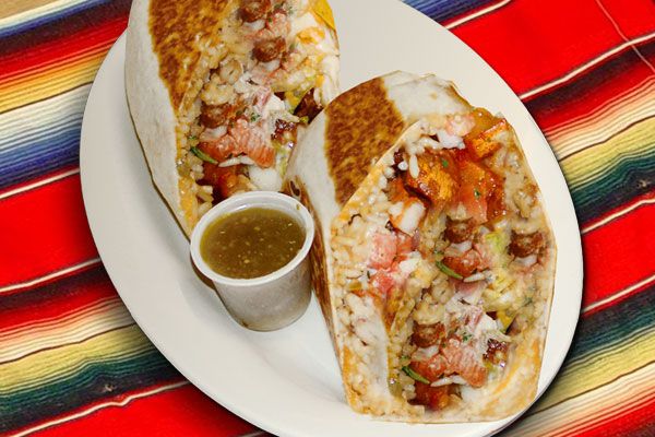 A burrito, wrapped in foil and sliced in half, sits on a white plate on a tablecloth with brightly colored stripes.