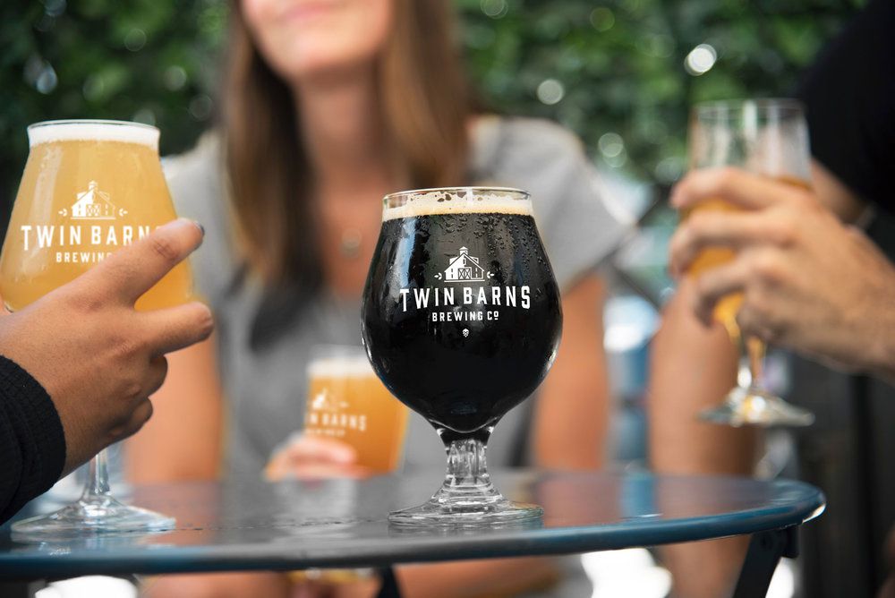 At Twin Barns Brewing Company, three people hoist full beer glasses in the background, while a full tulip glass of dark beer sits centered on a table