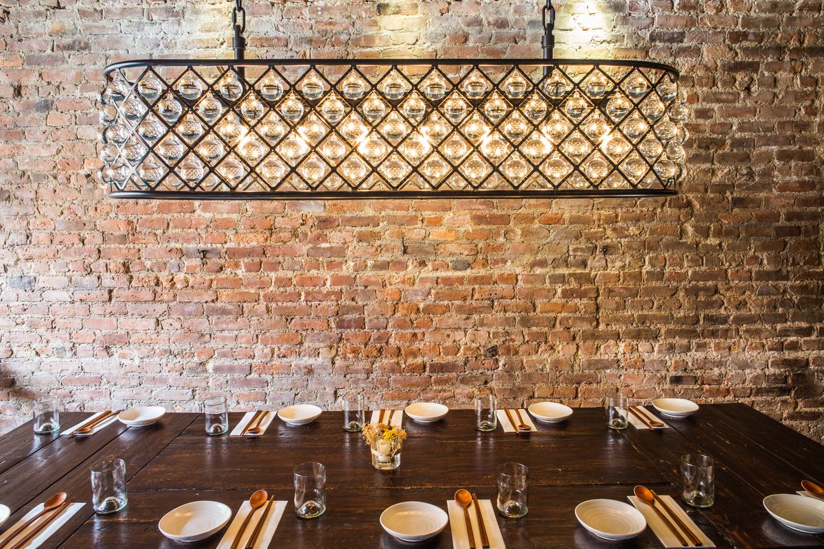 A large light fixture hangs over a dark wooden table set with plates and glasses. A red brick wall is in the background.