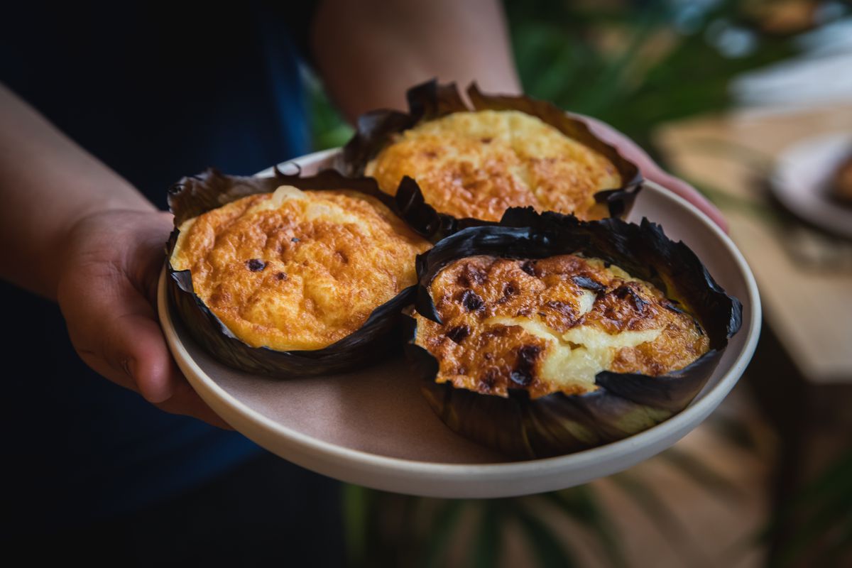 A ceramic plate with three bibingka, wrapped in a leaf, with a golden crust and a chewy-looking, rich middle.