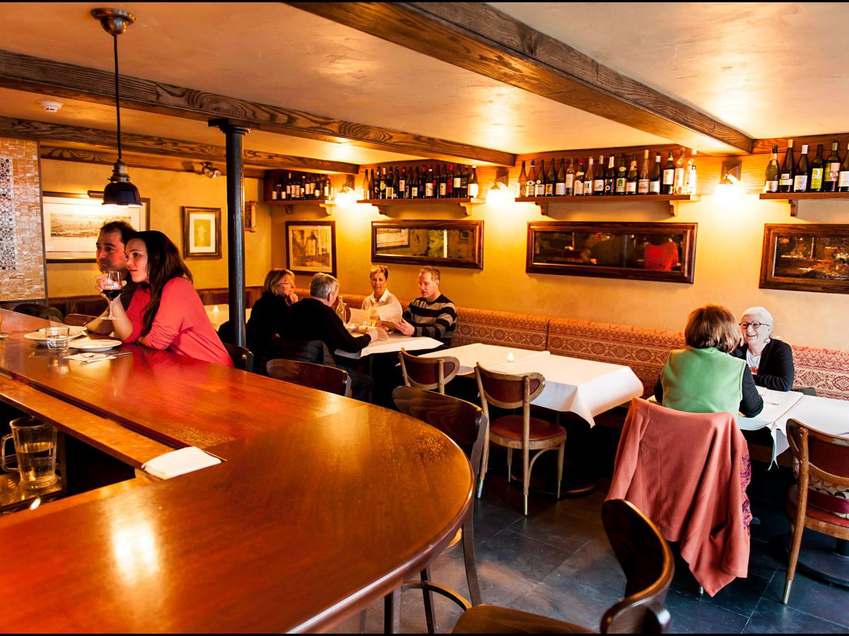 Diners sit at tables along a wine-rack lined wall and at a bar, in a low-lit tavern like room