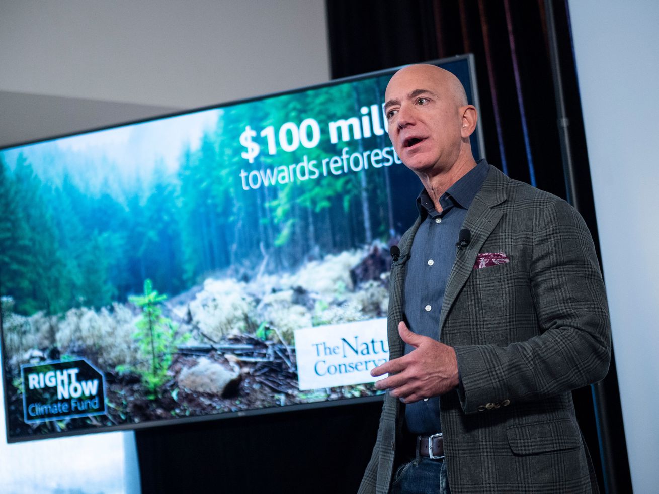Amazon founder Jeff Bezos speaks onstage in front of a screen showing a wooded landscape and the words “$100 million.”