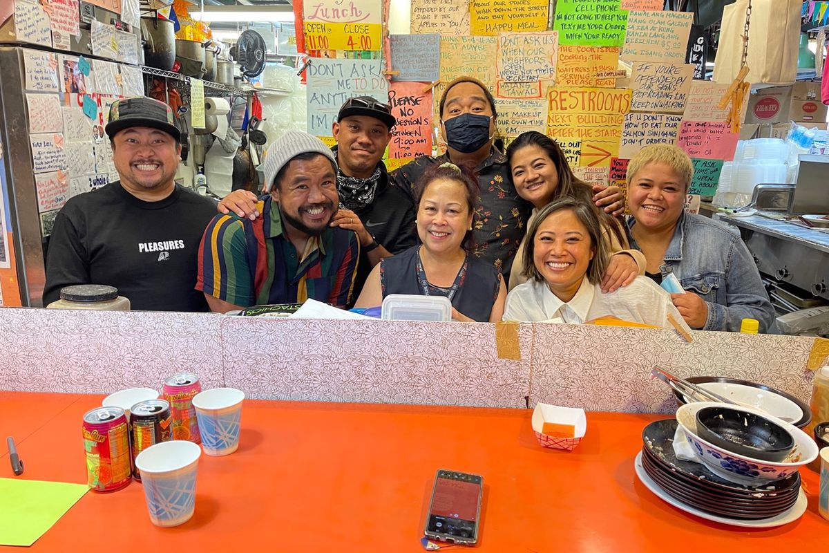 A group of smiling brown-skinned people stand together taking a mirror selfie. Hand-written signs and notices are posted on the wall behind them.